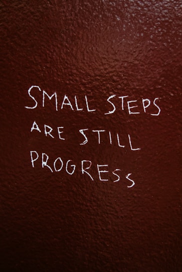 Text is written on a textured, dark brown wall. The phrase reads, 'Small steps are still progress' in white, slightly uneven handwriting.