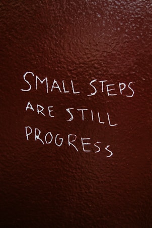 Text is written on a textured, dark brown wall. The phrase reads, 'Small steps are still progress' in white, slightly uneven handwriting.