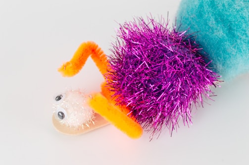 A whimsical craft creation features a combination of colorful materials including a fuzzy blue pom-pom, a bright pink tinsel pom-pom, a white glittery pom-pom with googly eyes, and arms made from orange pipe cleaners. The elements are arranged to resemble a playful creature.