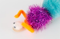 A whimsical craft creation features a combination of colorful materials including a fuzzy blue pom-pom, a bright pink tinsel pom-pom, a white glittery pom-pom with googly eyes, and arms made from orange pipe cleaners. The elements are arranged to resemble a playful creature.