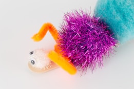 A whimsical craft creation features a combination of colorful materials including a fuzzy blue pom-pom, a bright pink tinsel pom-pom, a white glittery pom-pom with googly eyes, and arms made from orange pipe cleaners. The elements are arranged to resemble a playful creature.