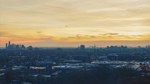 The image captures an expansive cityscape under a dramatic sunset. The skyline features a mix of modern skyscrapers and urban buildings. Construction cranes are visible, indicating ongoing development. The sky is filled with vibrant oranges, yellows, and soft clouds.