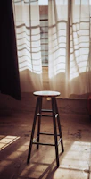 A rustic wooden bench stool placed beside a sunlit window with soft cushions.