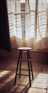 A stylish wooden bench stool placed beside a sunlit window with a soft throw blanket.