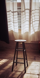 A handcrafted wooden stool with a natural finish placed beside a large window with sunlight streaming in.