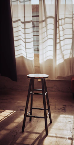 A rustic wooden bench stool placed beside a sunlit window with soft cushions.