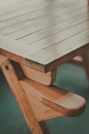 A wooden table with visible grain and a handcrafted design, showcasing a natural finish and sturdy construction. The photo captures a close-up angle of the table's edge and legs, highlighting its rustic charm.