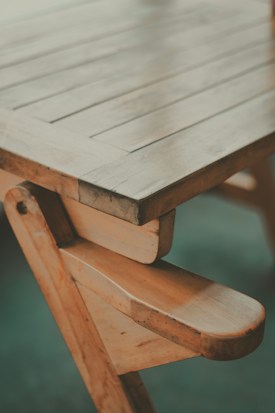 A wooden table with visible grain and a handcrafted design, showcasing a natural finish and sturdy construction. The photo captures a close-up angle of the table's edge and legs, highlighting its rustic charm.