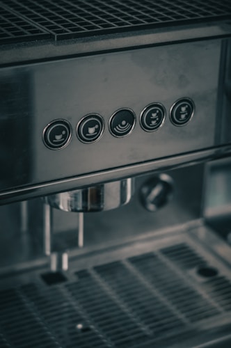 A close-up view of a stainless steel coffee machine with five circular buttons on the front panel. The buttons have icons indicating different coffee options. The machine has a grid-like drip tray and a portafilter attached below.