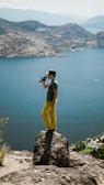 man in yellow shorts holding black dslr camera standing on brown rock near body of water