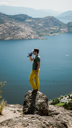 man in yellow shorts holding black dslr camera standing on brown rock near body of water