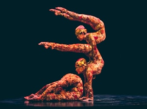 Two performers are engaged in a complex acrobatic pose on a dark stage. They are wearing multicolored, intricate bodysuits that blend red, pink, and gold hues. The performer's positions demonstrate strength and balance.