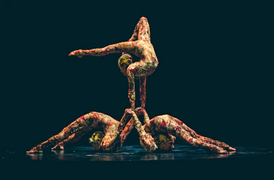 Three performers in colorful, patterned bodysuits are engaged in a balanced acrobatic act. The composition involves two individuals supporting a third performer, who extends horizontally above them. The background is dark, emphasizing the vibrant costumes and the intricate pose.