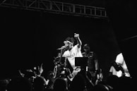 Black and white photograph capturing a live music performance. A performer is energetically singing on stage, wearing sunglasses and a t-shirt, holding a microphone. The audience in the foreground is capturing the moment with their phones. Stage lighting and equipment are visible, contributing to a dynamic atmosphere.