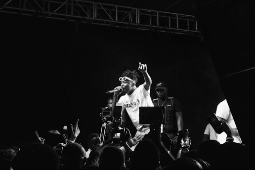 Black and white photograph capturing a live music performance. A performer is energetically singing on stage, wearing sunglasses and a t-shirt, holding a microphone. The audience in the foreground is capturing the moment with their phones. Stage lighting and equipment are visible, contributing to a dynamic atmosphere.