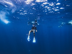 A person wearing snorkeling gear, including fins and a mask, is underwater in a vast blue ocean. Sunlight creates a shimmering effect on the surface above, casting light streaks through the water. Other divers are visible in the background, exploring the aquatic environment.