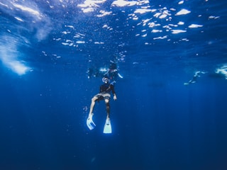 A person wearing snorkeling gear, including fins and a mask, is underwater in a vast blue ocean. Sunlight creates a shimmering effect on the surface above, casting light streaks through the water. Other divers are visible in the background, exploring the aquatic environment.