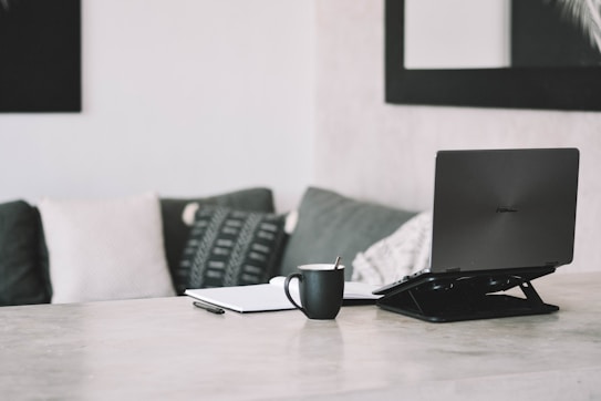 A modern, minimalist workspace featuring a laptop on a wooden table, accompanied by a notebook, a pen, and a black coffee mug. The background includes a sofa with patterned and plain cushions, adding a touch of coziness. The setting is complemented by a large framed mirror on the wall, enhancing the room's spaciousness.