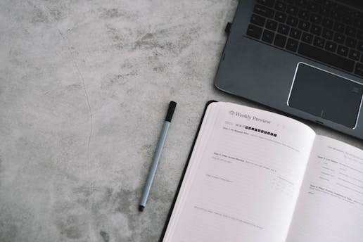 An open planner with the heading 'Weekly Preview' next to a laptop on a gray textured surface. A pen lies between the planner and the laptop.
