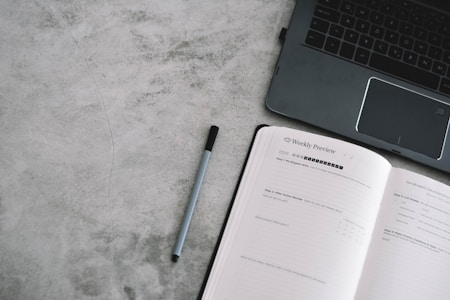 An open planner with the heading 'Weekly Preview' next to a laptop on a gray textured surface. A pen lies between the planner and the laptop.