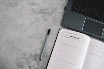 An open planner with the heading 'Weekly Preview' next to a laptop on a gray textured surface. A pen lies between the planner and the laptop.