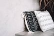Two decorative pillows rest against a neutral-toned concrete wall. The pillow on the left is black with white geometric patterns and tassels, while the one on the right is white with black arrow patterns. A few dried leaves are visible in the top right corner.