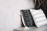 Two decorative pillows rest against a neutral-toned concrete wall. The pillow on the left is black with white geometric patterns and tassels, while the one on the right is white with black arrow patterns. A few dried leaves are visible in the top right corner.