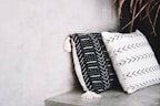 Two decorative pillows rest against a neutral-toned concrete wall. The pillow on the left is black with white geometric patterns and tassels, while the one on the right is white with black arrow patterns. A few dried leaves are visible in the top right corner.