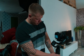 A person with tattoos on their left arm operates a camera mounted on a gimbal inside a modern living room. The area has a white wall, a TV on a stand, and various decorative elements like potted plants and a patterned wallpaper.