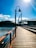 A wooden boardwalk stretches alongside a body of water under a clear blue sky. In the background, a modern bridge structure with people walking across it extends over the water. A street lamp stands prominently on the boardwalk, and trees line the opposite shore.