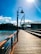 A wooden boardwalk stretches alongside a body of water under a clear blue sky. In the background, a modern bridge structure with people walking across it extends over the water. A street lamp stands prominently on the boardwalk, and trees line the opposite shore.