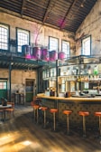 A stylish industrial-style bar with high ceilings and vintage decor. There are large windows allowing natural light to flood the space, highlighting the leather bar stools and a sleek, curved bar counter. The upper level features large brewing tanks, and exposed brick walls add to the rustic charm.