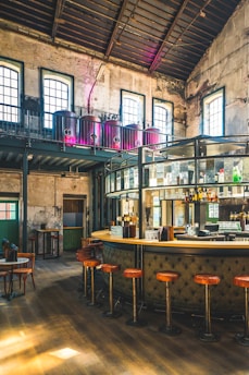 A stylish industrial-style bar with high ceilings and vintage decor. There are large windows allowing natural light to flood the space, highlighting the leather bar stools and a sleek, curved bar counter. The upper level features large brewing tanks, and exposed brick walls add to the rustic charm.