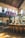 A stylish industrial-style bar with high ceilings and vintage decor. There are large windows allowing natural light to flood the space, highlighting the leather bar stools and a sleek, curved bar counter. The upper level features large brewing tanks, and exposed brick walls add to the rustic charm.