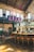 A stylish industrial-style bar with high ceilings and vintage decor. There are large windows allowing natural light to flood the space, highlighting the leather bar stools and a sleek, curved bar counter. The upper level features large brewing tanks, and exposed brick walls add to the rustic charm.