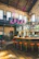 A stylish industrial-style bar with high ceilings and vintage decor. There are large windows allowing natural light to flood the space, highlighting the leather bar stools and a sleek, curved bar counter. The upper level features large brewing tanks, and exposed brick walls add to the rustic charm.