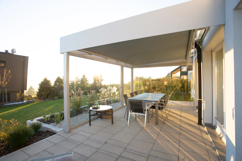 The image depicts a modern patio with a large glass-enclosed table surrounded by six chairs, all facing towards an expansive view of trees and houses beyond. The patio is made of concrete and features a white roof that provides shade from the sun. A small garden area can be seen in front of the patio, adding to the overall aesthetic appeal of the space.