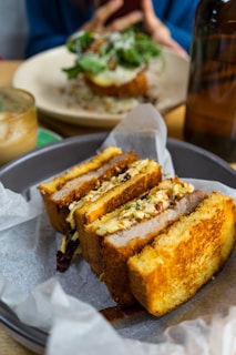 A close-up of a neatly arranged sandwich with crispy, golden-brown bread and a filling of meat and vegetables. The sandwich is displayed in a round, dark plate lined with parchment paper. In the background, there is another dish and a blurred person capturing a photo with a smartphone.