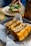 A close-up of a neatly arranged sandwich with crispy, golden-brown bread and a filling of meat and vegetables. The sandwich is displayed in a round, dark plate lined with parchment paper. In the background, there is another dish and a blurred person capturing a photo with a smartphone.