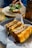 A close-up of a neatly arranged sandwich with crispy, golden-brown bread and a filling of meat and vegetables. The sandwich is displayed in a round, dark plate lined with parchment paper. In the background, there is another dish and a blurred person capturing a photo with a smartphone.