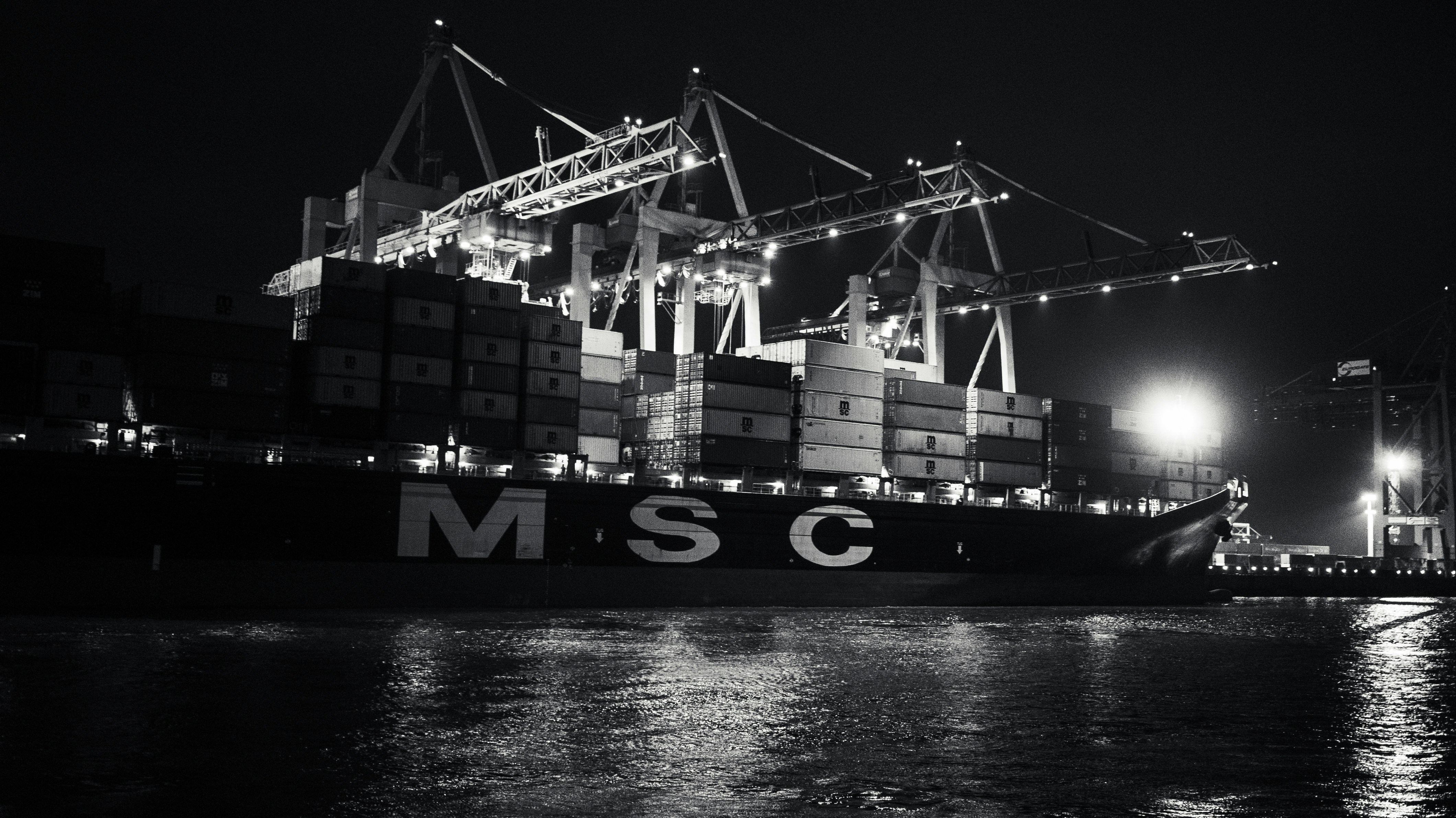 Port operations at night, bright floodlights illuminating container cranes, dark sky, active cargo loading operations