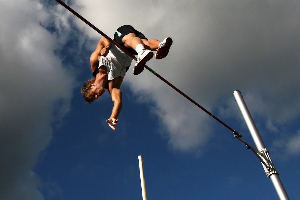 man in white tank top and black shorts hanging on black metal bar under blue sky