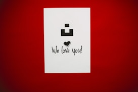A white card with a black geometric symbol at the top, a heart graphic in the middle, and the phrase 'We love you!' in an expressive font at the bottom, all set against a deep red background.