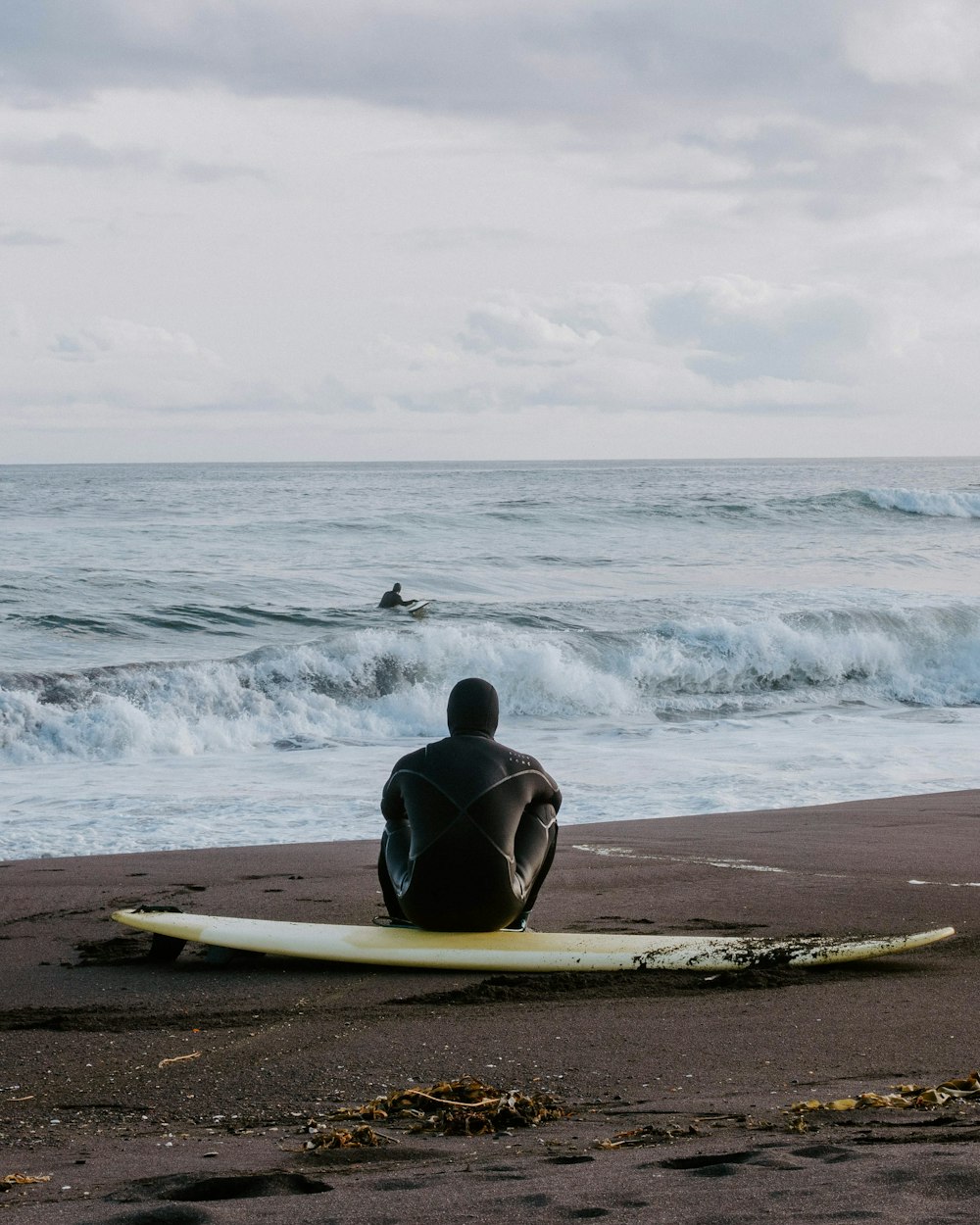 uomo in muta umida nera che si siede sulla tavola da surf bianca sulla spiaggia durante il giorno