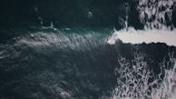 Aerial view of a surfer riding a wave in the ocean, with foamy white water contrasting against dark green and blue hues of the sea.