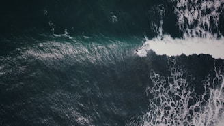 Aerial view of a surfer riding a wave in the ocean, with foamy white water contrasting against dark green and blue hues of the sea.
