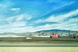 A bright red truck with a flatbed trailer is parked on a grassy area, with a backdrop of rolling hills and mountains under a blue sky with scattered clouds. A highway runs parallel in the foreground.