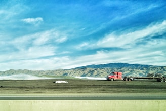 A bright red truck with a flatbed trailer is parked on a grassy area, with a backdrop of rolling hills and mountains under a blue sky with scattered clouds. A highway runs parallel in the foreground.