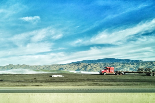 A bright red truck with a flatbed trailer is parked on a grassy area, with a backdrop of rolling hills and mountains under a blue sky with scattered clouds. A highway runs parallel in the foreground.
