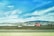 A bright red truck with a flatbed trailer is parked on a grassy area, with a backdrop of rolling hills and mountains under a blue sky with scattered clouds. A highway runs parallel in the foreground.